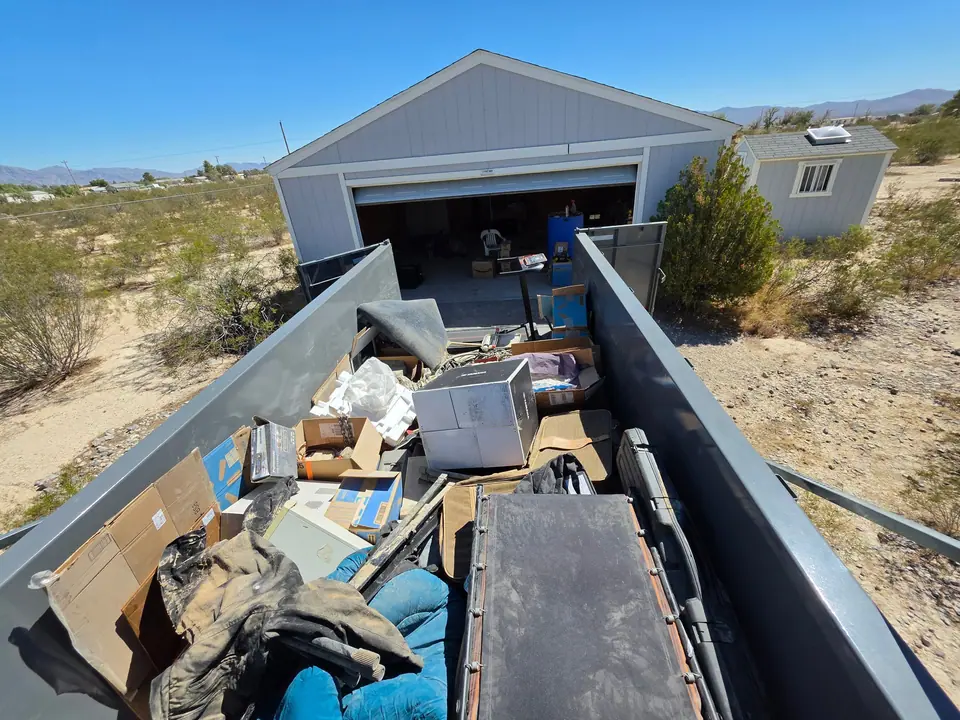 Dump trailer loaded with boxes and household items outside an open garage on a desert lot