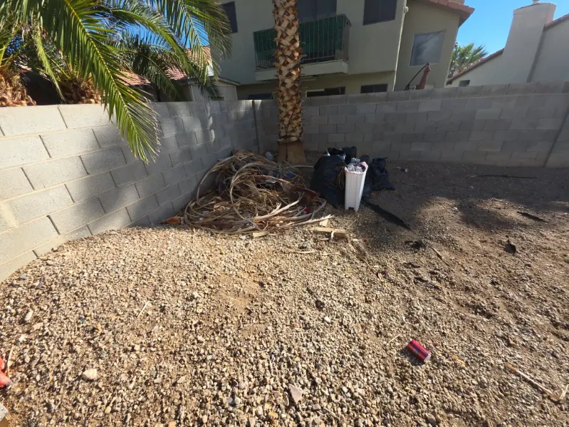 Backyard gravel with a pile of dead palm fronds and bagged trash against a block wall