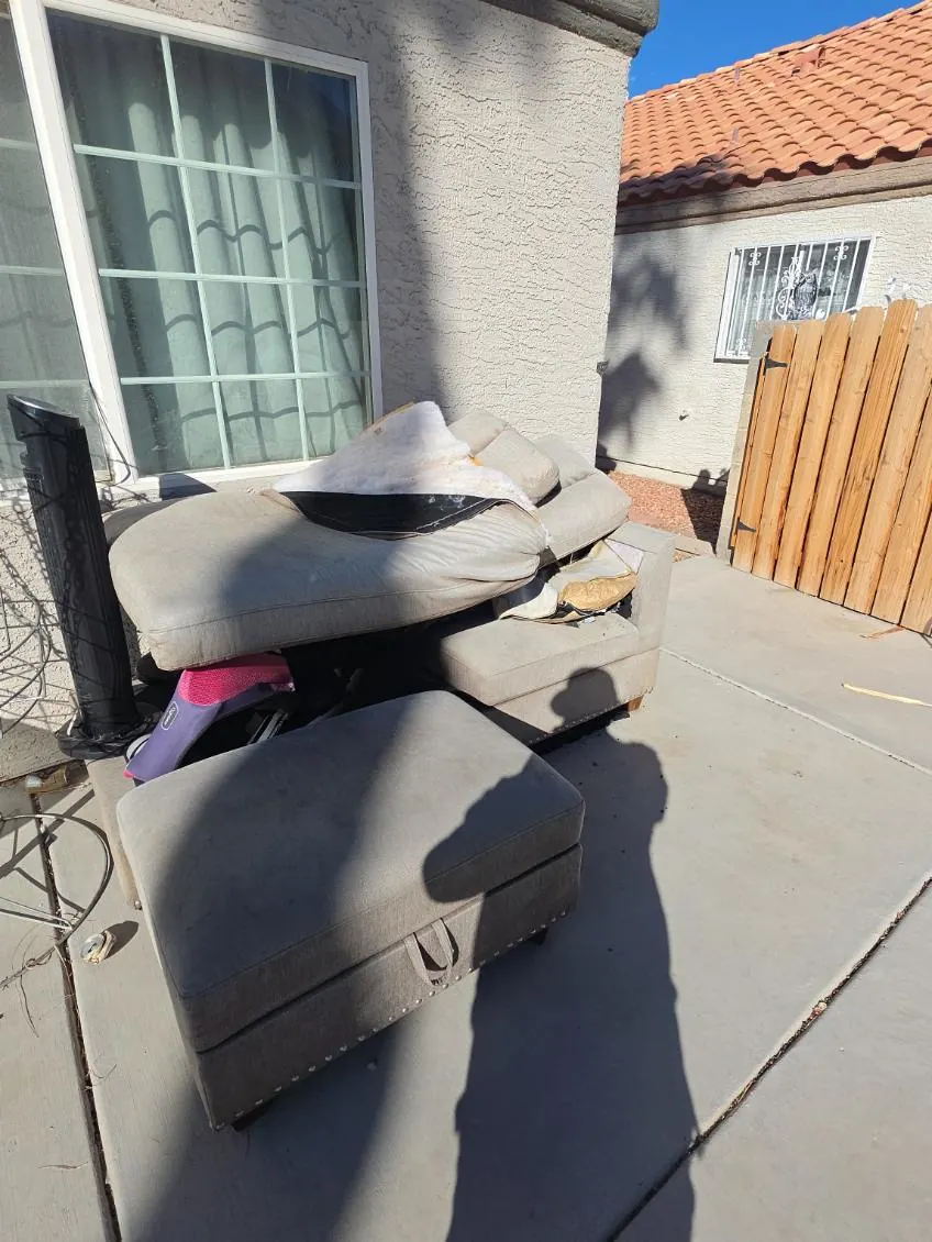 Worn grey sectional and ottoman piled on a back patio beside a stucco house